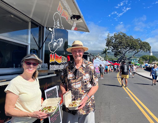 two people standing outside beside a black food truck, holding food